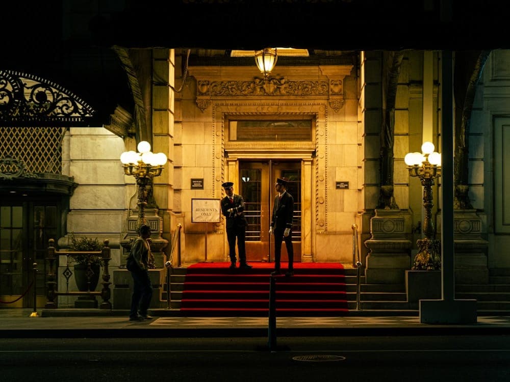 Entrada iluminada de um edifício à noite, com dois porteiros uniformizados de plantão.